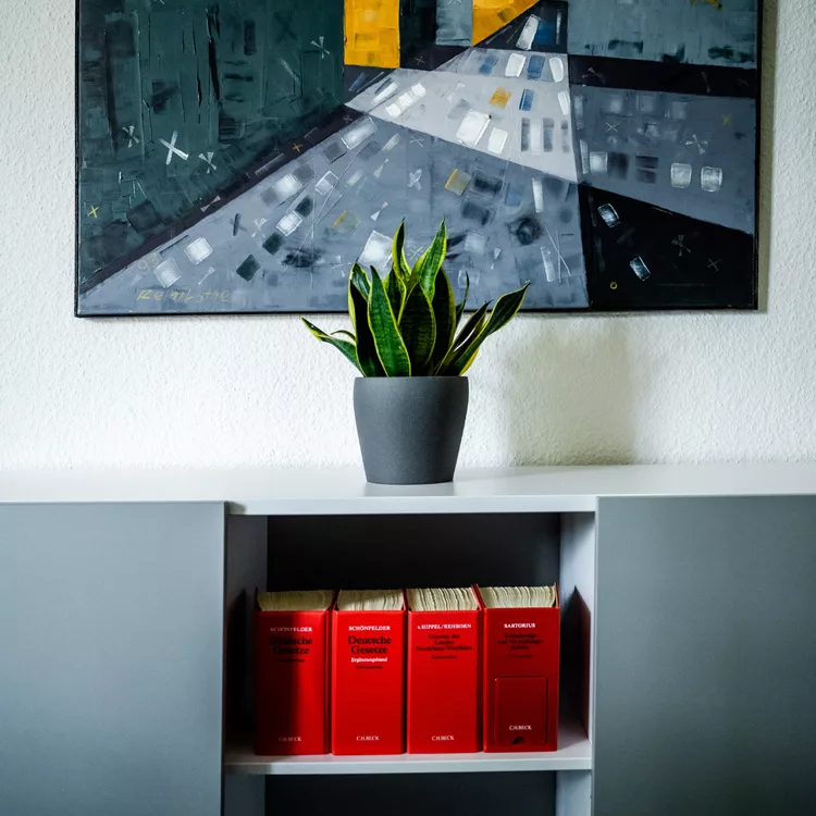 Auf einem Regal steht ein eingetopfte Pflanze, im Hintergrund ist ein großes modernes Gemälde, auf dem Hochhäuser zu sehen sind